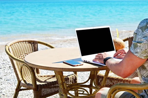 Young man working on his laptop at the beach at tropical destination. Office on the beach concept.