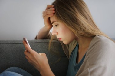 Telefonunu tutan gri tekstil kanepede oturan depresif yüz ifadesi ile güzel genç kadının portresi. Siber zorbalık kurbanı kavramı. Odasında üzgün bir kadın var. Arka plan, kopyalama alanı.