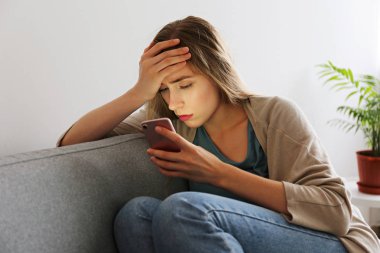 Telefonunu tutan gri tekstil kanepede oturan depresif yüz ifadesi ile güzel genç kadının portresi. Siber zorbalık kurbanı kavramı. Odasında üzgün bir kadın var. Arka plan, kopyalama alanı.