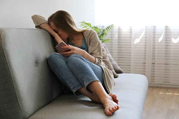 Ritratto di giovane donna depressa che nasconde il viso seduta sul divano tessile grigio con il telefono in mano. Il concetto di vittima di bullismo informatico. Donna triste nella sua stanza. Sfondo, spazio di copia . — Foto Stock