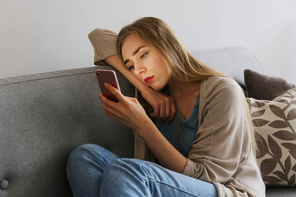 Ritratto di bella giovane donna con espressione facciale depressa seduta sul divano tessile grigio con il suo telefono in mano. Il concetto di vittima di bullismo informatico. Donna triste nella sua stanza. Sfondo, spazio di copia . — Foto Stock