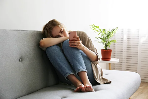 Porträtt av vacker ung kvinna med deprimerad ansiktsuttryck sitter på grå textil soffa hålla hennes telefon. Cybermobbning offer koncept. Ledsen kvinna i hennes rum. Bakgrund, kopiera utrymme. — Stockfoto