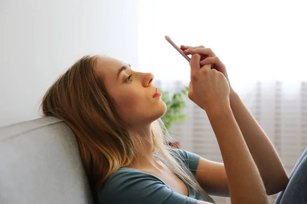 Ritratto di bella giovane donna con espressione facciale depressa seduta sul divano tessile grigio con il suo telefono in mano. Il concetto di vittima di bullismo informatico. Donna triste nella sua stanza. Sfondo, spazio di copia . — Foto Stock