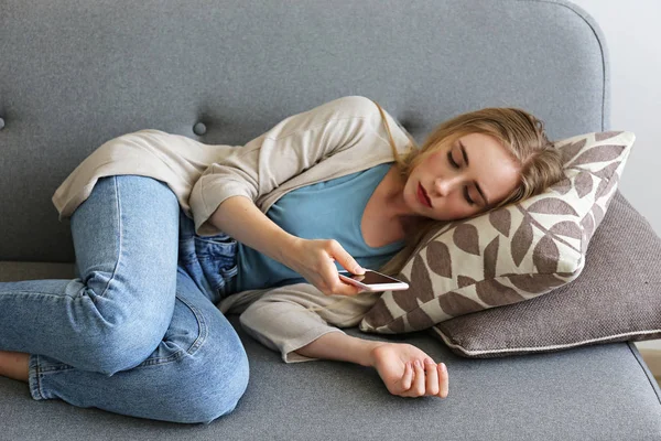 Ritratto di bella giovane donna con espressione facciale depressa seduta sul divano tessile grigio con il suo telefono in mano. Il concetto di vittima di bullismo informatico. Donna triste nella sua stanza. Sfondo, spazio di copia . — Foto Stock