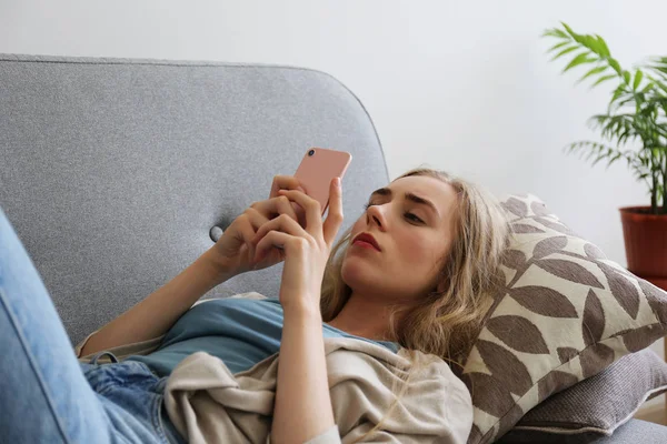 Telefonunu tutan gri tekstil kanepede oturan depresif yüz ifadesi ile güzel genç kadının portresi. Siber zorbalık kurbanı kavramı. Odasında üzgün bir kadın var. Arka plan, kopyalama alanı. — Stok fotoğraf