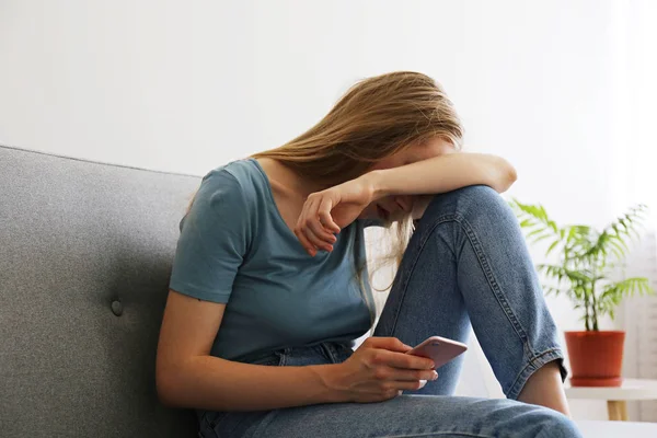 Portrait of depressed young woman hiding her face sitting on grey textile couch holding the phone. Cyber bullying victim concept. Sad female in her room. Background, copy space. — Stock Photo, Image