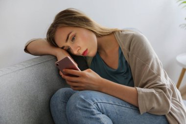 Telefonunu tutan gri tekstil kanepede oturan depresif yüz ifadesi ile güzel genç kadının portresi. Siber zorbalık kurbanı kavramı. Odasında üzgün bir kadın var. Arka plan, kopyalama alanı.