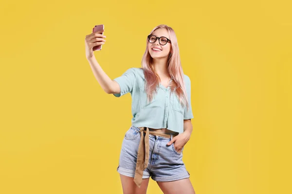Junge schöne blonde Frau mit pinkfarbenem Haartoner posiert vor grellem, buntem Hintergrund und hält ihr Smartphone in der Hand. Porträt eines weiblichen Teenie-Models, das Emotionen zeigt. Nahaufnahme, Kopierraum. — Stockfoto