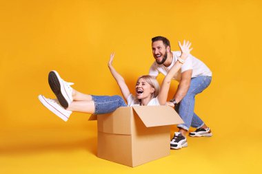 Young couple in matching outfits having fun moving boxes, isolated on yellow. clipart