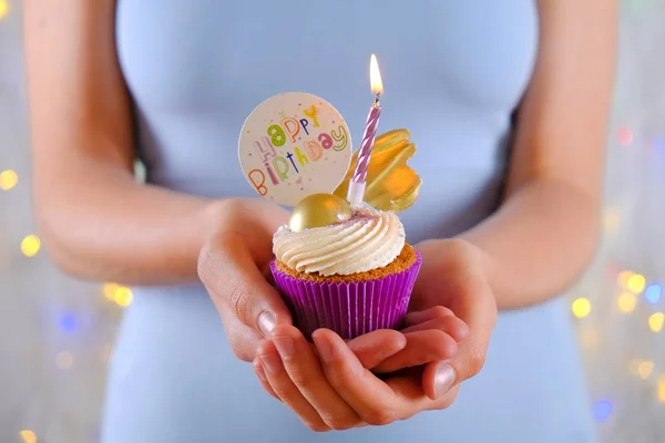 Kvinna som innehar Grattis på födelsedagen cupcake med brinnande ljus i lila wrap, grädde ost glasyr virvel och gyllene choklad dekoration på vitt trä texturerat bord. Närbild, kopiera utrymme, bakgrund. — Stockfoto