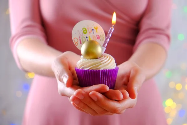 Mujer sosteniendo cupcake feliz cumpleaños con vela ardiente en envoltura púrpura, crema de queso glaseado remolino y decoración de chocolate dorado en la mesa de textura de madera blanca. Cerrar, copiar espacio, fondo . —  Fotos de Stock