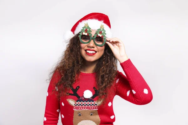 Studio portret van jonge vrouw met donkere huid en lange krullend haar dragen strakke kerstman hoed en kerst outfit. Lelijk trui concept. Close up, kopieerruimte voor tekst, geïsoleerde achtergrond. — Stockfoto