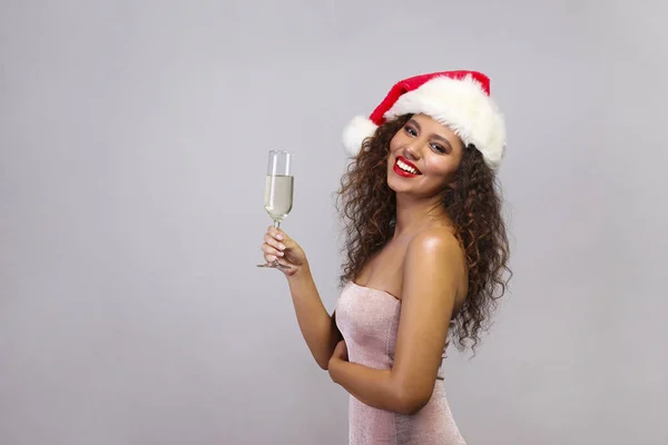 Studio ritratto di giovane donna con pelle scura e lunghi capelli ricci indossa abito stretto sexy e cappello di Babbo Natale, tenendo bicchiere di champagne. Chiudi, copia spazio per testo, sfondo isolato . — Foto Stock