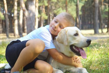Evcil arkadaşı Labrador Retriever ile ormanda takılan sevimli sarışın çocuğun portresi. Ufaklık altı aylık köpeğe sarılıyor, parkta yürüyor. Arkaplan, kopyala boşluk, kapat.