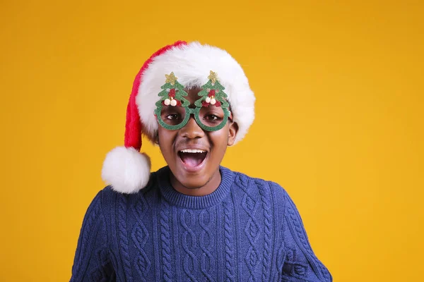 Афроамериканець, одягнений в стильну сукню, посміхається у капелюсі з подарунком на жовтому тлі studio. темношкірий Санта Клаус вітає веселі кристали. — стокове фото