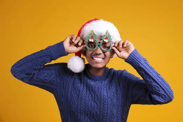 Афроамериканець, одягнений в стильну сукню, посміхається у капелюсі з подарунком на жовтому тлі studio. темношкірий Санта Клаус вітає веселі кристали. — стокове фото
