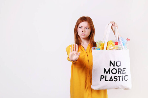 Emotional redhead woman holding eco friendly shopping bag with no more plastic text, full of harmful products, bottles and bags. Zero waste concept. Happy female sorting, separating garabage.