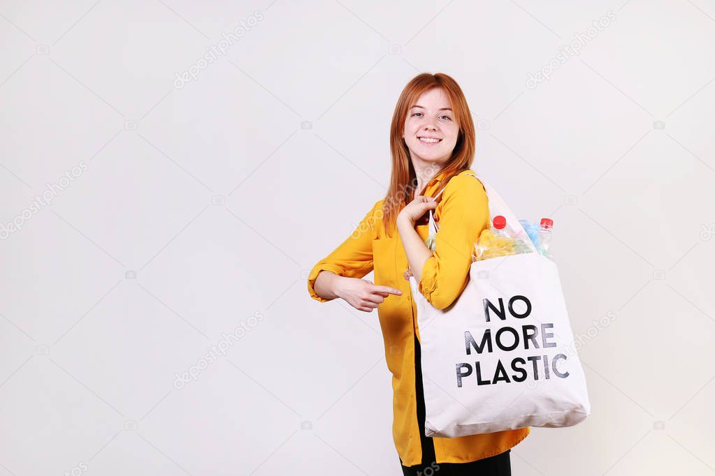 Emotional redhead woman holding eco friendly shopping bag with no more plastic text, full of harmful products, bottles and bags. Zero waste concept. Happy female sorting, separating garabage.