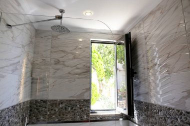 Banyo açık bir pencereden içeri doğal ışık ile dolu. Duş başlığı akan suyla tavana monte edildi. Modern çerçevesiz cam panel, mermer desenli fayans. Boşluğu kopyala, arkaplan.