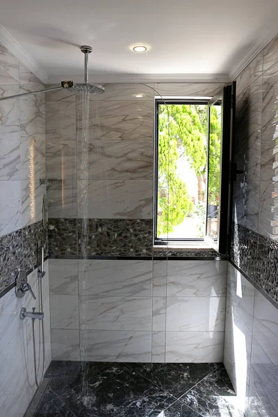窓から自然光が差し込む浴室 シャワーヘッドはランニングウォーターで天井に取り付けられます 現代的なフレームのない固定ガラスパネル 大理石のテクスチャタイル コピースペース — ストック写真
