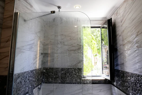 窓から自然光が差し込む浴室 シャワーヘッドはランニングウォーターで天井に取り付けられます 現代的なフレームのない固定ガラスパネル 大理石のテクスチャタイル コピースペース — ストック写真