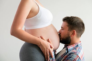 Kareli gömlekli sakallı bir adamın portresi. Karısının hamile karnını yalıtılmış beyaz arka planda öpüyor. Doğum öncesi dönem konsepti. Genç bir aile yeni bir çocuk bekliyor. Yakın çekim..