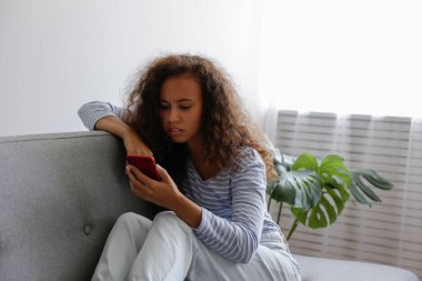 Yüzünde depresif bir ifade olan, gri tekstil kanepesinde oturan ve telefonunu tutan güzel zenci bir kadının portresi. Siber zorbalık mağduru konsepti. Odasında üzgün bir kadın var. Arkaplan, boşluğu kopyala.