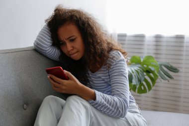 Yüzünde depresif bir ifade olan, gri tekstil kanepesinde oturan ve telefonunu tutan güzel zenci bir kadının portresi. Siber zorbalık mağduru konsepti. Odasında üzgün bir kadın var. Arkaplan, boşluğu kopyala.