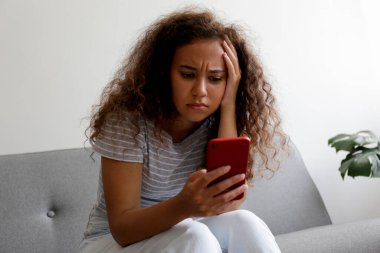 Yüzünde depresif bir ifade olan, gri tekstil kanepesinde oturan ve telefonunu tutan güzel zenci bir kadının portresi. Siber zorbalık mağduru konsepti. Odasında üzgün bir kadın var. Arkaplan, boşluğu kopyala.