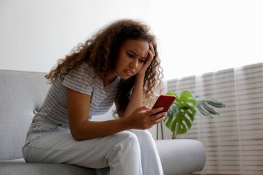 Yüzünde depresif bir ifade olan, gri tekstil kanepesinde oturan ve telefonunu tutan güzel zenci bir kadının portresi. Siber zorbalık mağduru konsepti. Odasında üzgün bir kadın var. Arkaplan, boşluğu kopyala.