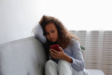Yüzünde depresif bir ifade olan, gri tekstil kanepesinde oturan ve telefonunu tutan güzel zenci bir kadının portresi. Siber zorbalık mağduru konsepti. Odasında üzgün bir kadın var. Arkaplan, boşluğu kopyala.