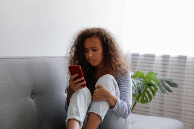 Yüzünde depresif bir ifade olan, gri tekstil kanepesinde oturan ve telefonunu tutan güzel zenci bir kadının portresi. Siber zorbalık mağduru konsepti. Odasında üzgün bir kadın var. Arkaplan, boşluğu kopyala.
