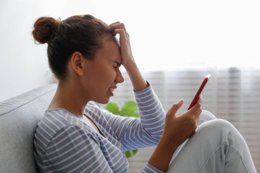 Yüzünde depresif bir ifade olan, gri tekstil kanepesinde oturan ve telefonunu tutan güzel zenci bir kadının portresi. Siber zorbalık mağduru konsepti. Odasında üzgün bir kadın var. Arkaplan, boşluğu kopyala.