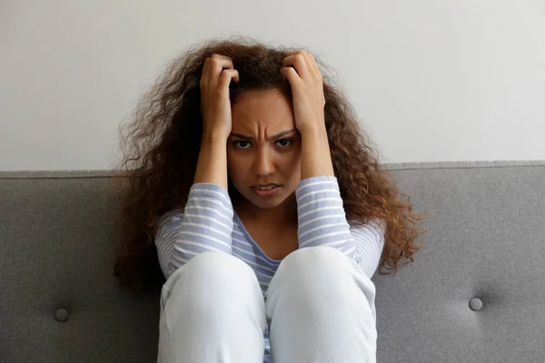 Portrait of young beautiful black woman with depressed facial expression sitting on the couch touching her temples. Female in physical and emotional pain. Sad girl in her room. Background, copy space.