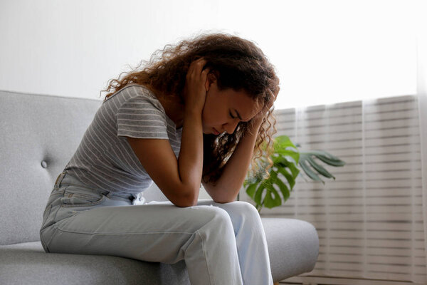Портрет молодой красивой черной женщины с депрессивным выражением лица, сидящей на диване и трогающей виски. Женщина в физической и эмоциональной боли. Грустная девушка в своей комнате. Фон, копировальное пространство.