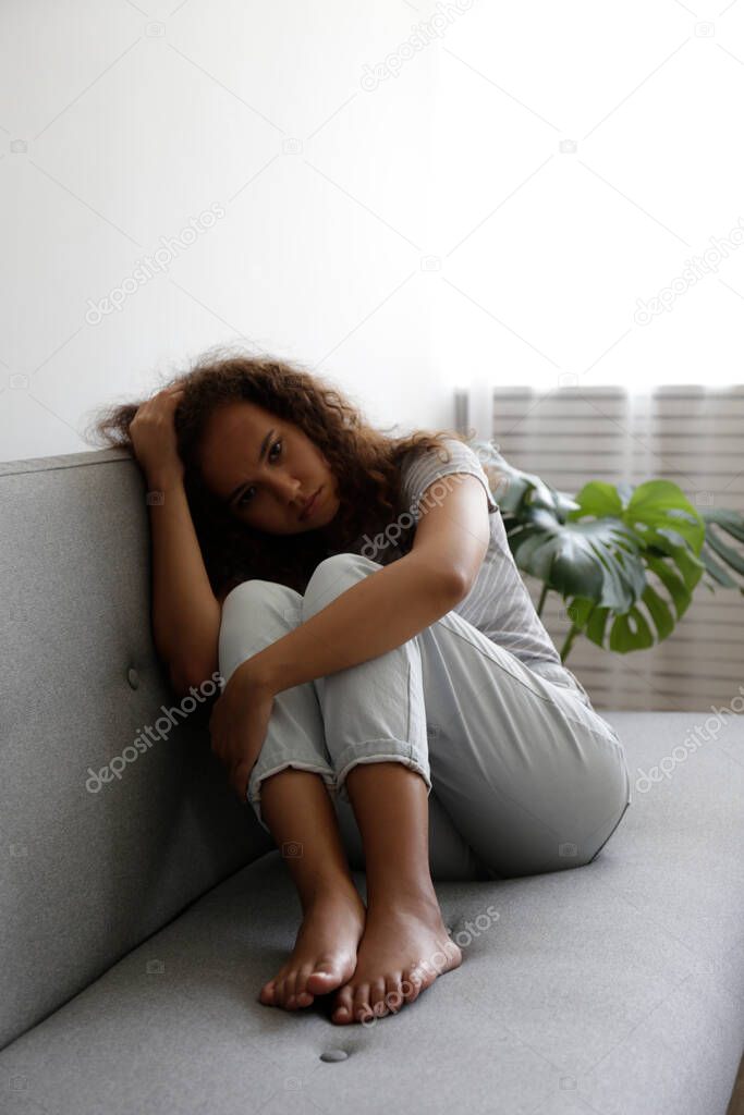 Portrait of young beautiful black woman with depressed facial expression sitting on the couch touching her temples. Female in physical and emotional pain. Sad girl in her room. Background, copy space.