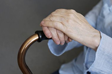 Huzurevinde oturan yaşlı bir kadın. Elinde kırış kırış bir elle yürüyen bir baston tutuyor. Bej hırka giyen yaşlı bir kadın, metal yardım çubuğu saplı çubuk, yakın çekim. İç arkaplan, kopyalama alanı