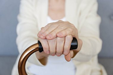 Huzurevinde oturan yaşlı bir kadın. Elinde kırış kırış bir elle yürüyen bir baston tutuyor. Yaşlı kadın bej hırka giyiyor, metal yardım çubuğu sapı yakın çekimde. İç arkaplan, kopyalama alanı