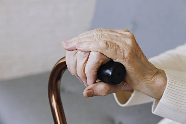 Elderly woman sitting in nursing home room holding walking quad cane with wrinkled hand. Old age lady wearing beige cardigan, metal aid stick handle bar close up. Interior background, copy space