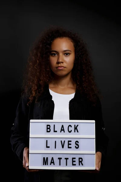 Portrait of young beautiful black woman standing in the dark with anti racist slogan. Protesting female holding a light box, black wall background. Close up, copy space.