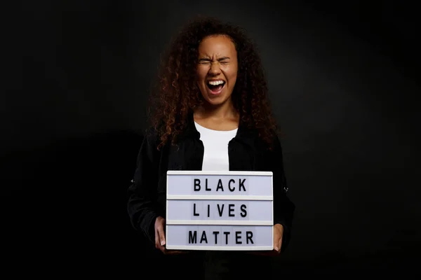 Portrait of young beautiful black woman standing in the dark with anti racist slogan. Protesting female holding a light box, black wall background. Close up, copy space.