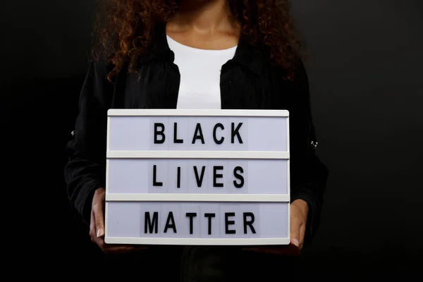 Portrait of young beautiful black woman standing in the dark with anti racist slogan. Protesting female holding a light box, black wall background. Close up, copy space.