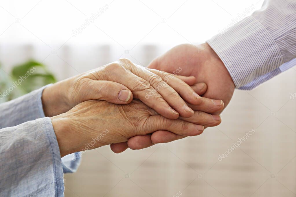 Mature female in elderly care facility gets help from hospital personnel nurse. Close up of aged wrinkled hands of senior woman reaching to a male doctor. Copy space, background