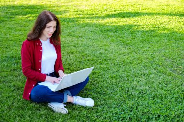 Genç ve güzel bir kadın yeşil çimlerin üzerinde dizüstü bilgisayarı üzerinde çalışıyor. Parktaki kablosuz sıcak noktaya bağlı. Çimenlikte bilgisayarlı esmer bir kadın internette geziniyor. Boşluğu kopyala, arkaplan.