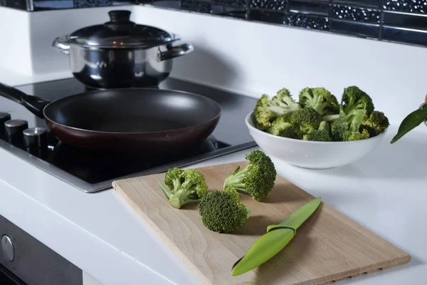 Tezgahın üzerinde beyaz seramik kasede pişmemiş yeşil çiğ brokoli yığını ve elektrikli sobanın üzerinde boş kızartma tavası. Vejetaryen diyeti. Kapat, boşluğu kopyala, arkaplan, üst görünüm.
