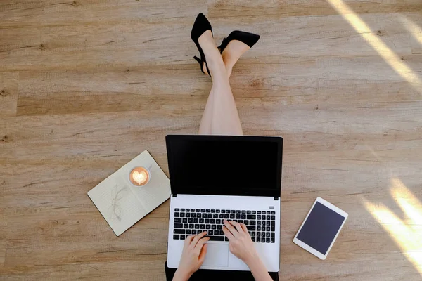 Siyah elbiseli genç bir kadının ahşap zeminde oturup dizüstü bilgisayarı üzerinde çalışmasının en iyi görüntüsü. Dişi ayağının kesilmiş hali. Kapat, kopyala boşluk, arkaplan.