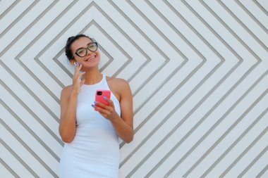 Arap kökenli, dar beyaz elbise giyen, akıllı telefonunu tutan, soyut geometrik desenlerle duvarın yanında duran genç, güzel bir kadın. Kapat, kopyala boşluk, arkaplan.