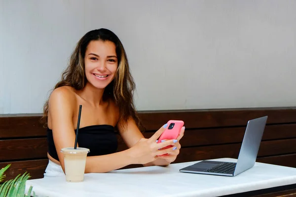 Young beautiful woman of Arabic ethnicity sitting by the city cafe table with laptop. Dark skinned female at coffee shop with iced latte. Close up, copy space, background.