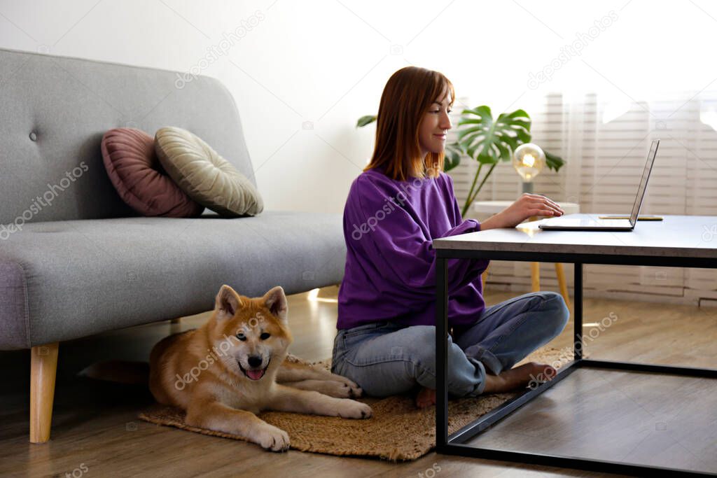 Portrait of a woman sitting on the floor, working on a laptop at home with her nine months old japanese akita inu pup. Happy female and funny big breed dog relaxing at home. Close up, copy space.