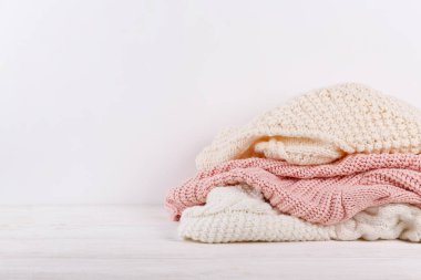 Beyaz ahşap masa üzerine yığılmış farklı örgü desenleri olan bir grup sıcak pastel renkli süveter, boş duvar arka planı. Sonbahar mevsimi örgü. Kapat, boşluğu kopyala.